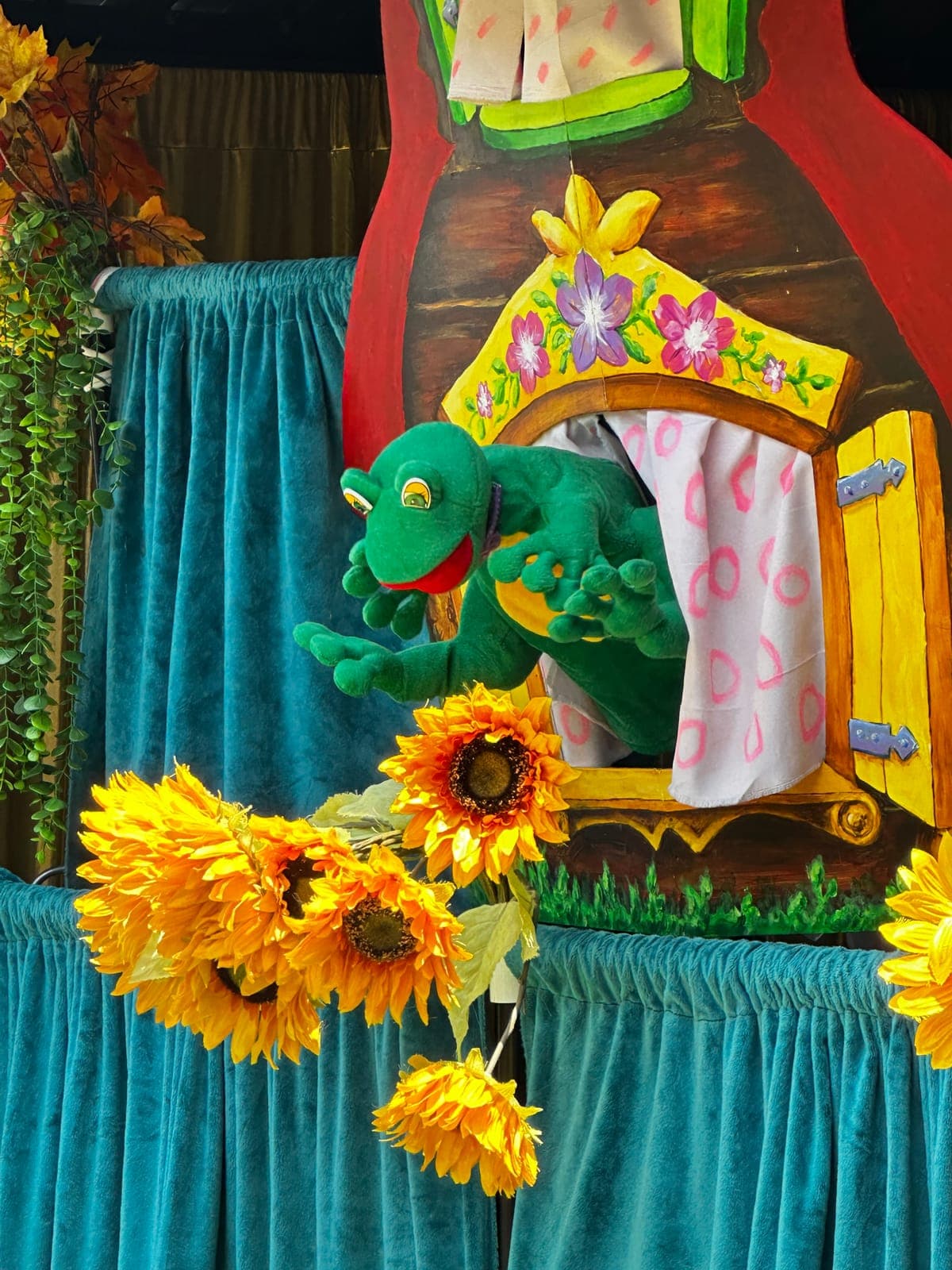 A cheerful green frog character leans from the flower-framed window of a painted storybook cottage.