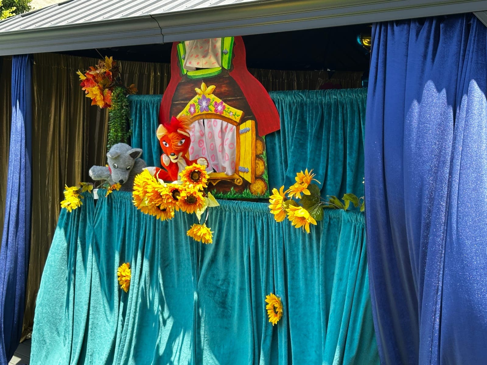 A whimsical outdoor stage with teal velvet drapes and sunflowers as the fox and wolf characters meet at a painted storybook cottage.