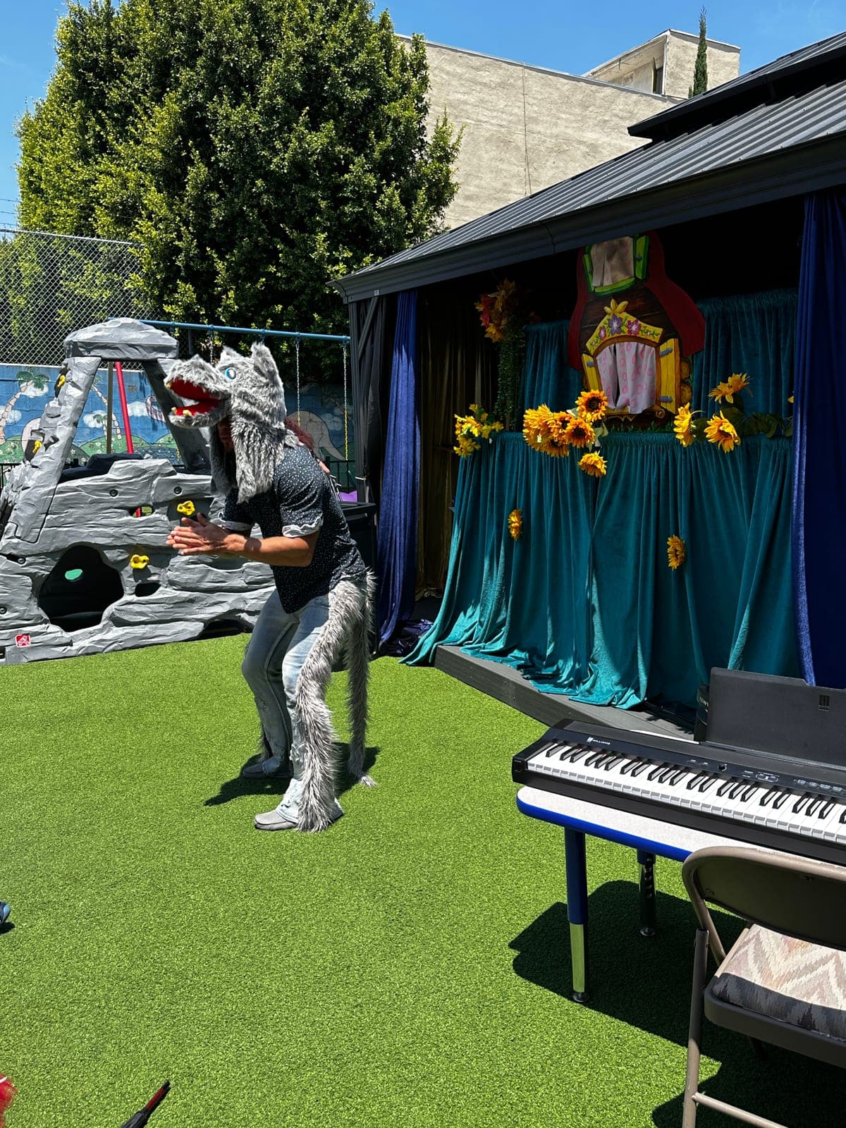 A performer in a full grey wolf costume acts out a scene before a decorated outdoor storybook stage.