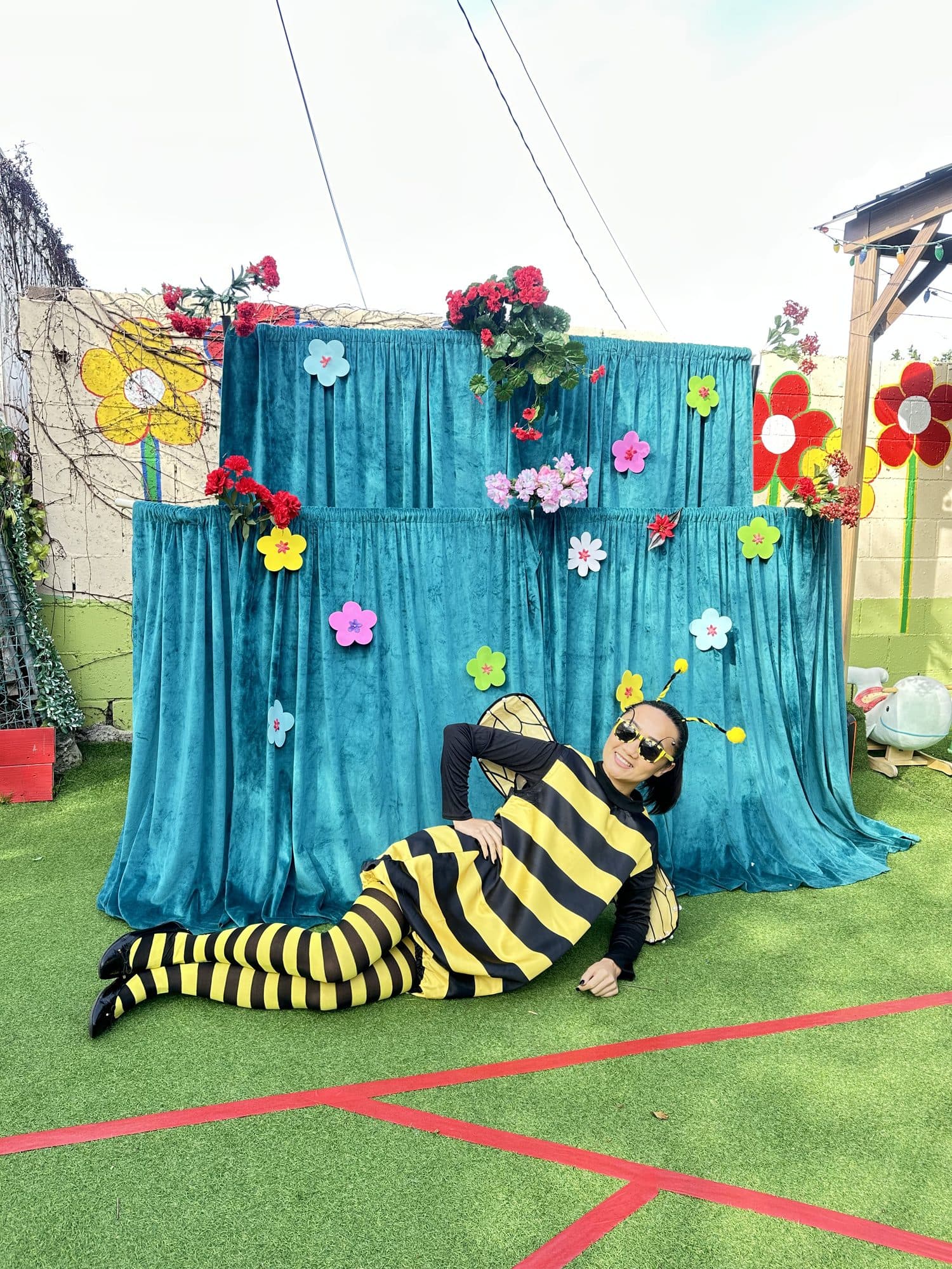 A performer in a black-and-yellow bee costume poses playfully before a flower-decked backdrop.