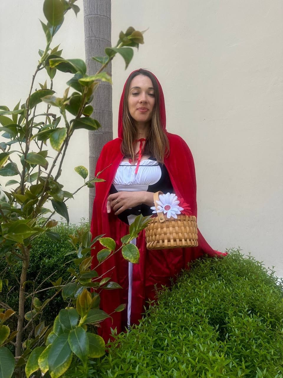 An actress in a red hooded cape holds a flower-trimmed basket outdoors.