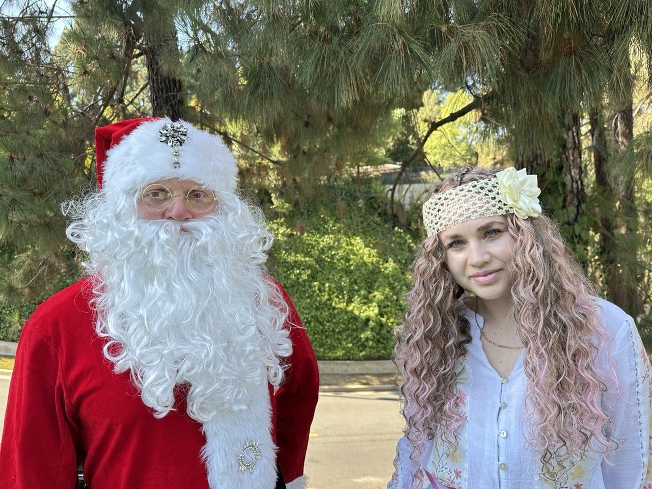 A performer dressed as Santa stands beside a smiling host among the pines.